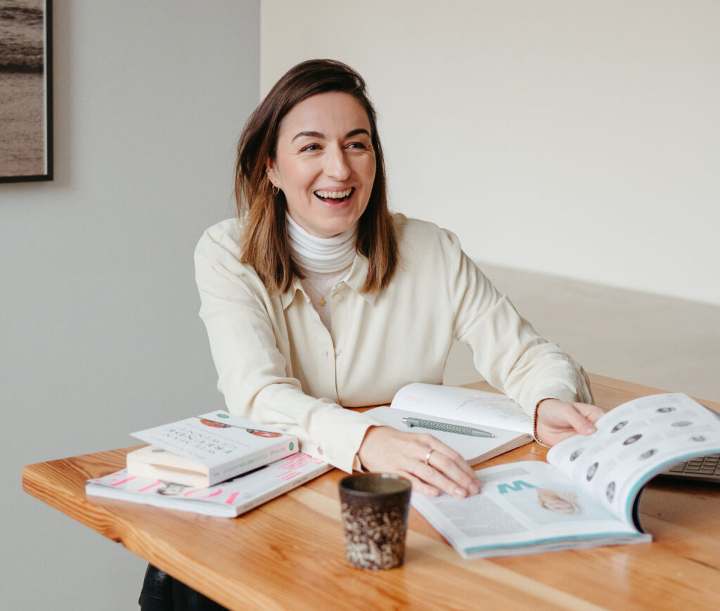 Henrike Redecker sitzt am Tisch, lacht und blättert in einem Magazin. Auf dem Tisch liegen Bücher und Notizen.