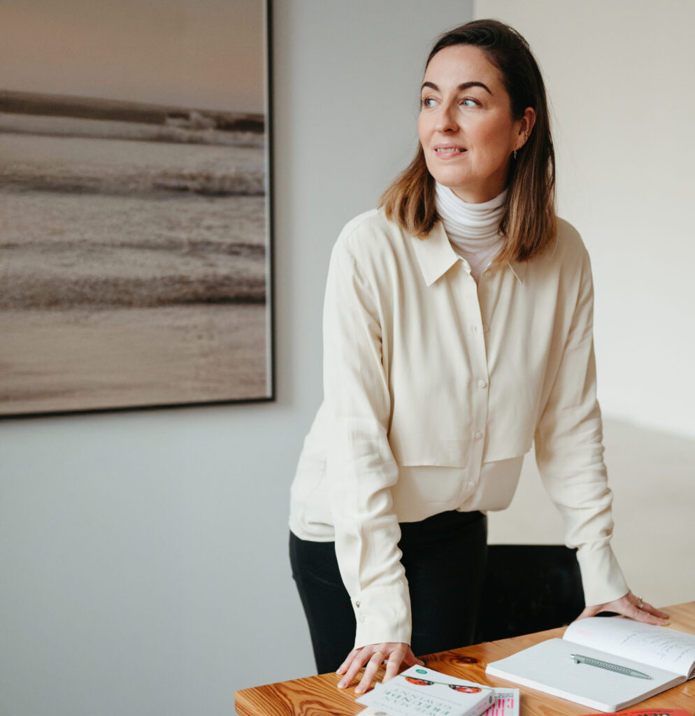 Henrike Redecker in cremefarbener Bluse lehnt am Holztisch und blickt nach rechts.
