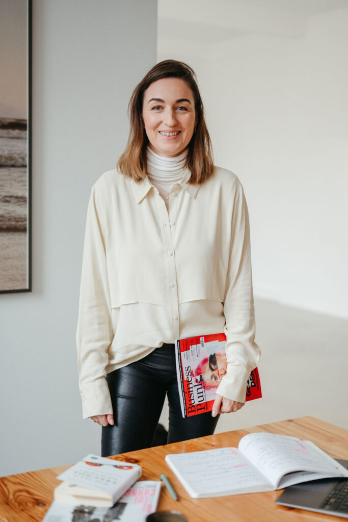 Henrike steht im Raum und hält das Magazin „Business Punk“; vor ihr liegt ein Tisch mit Notizbuch.