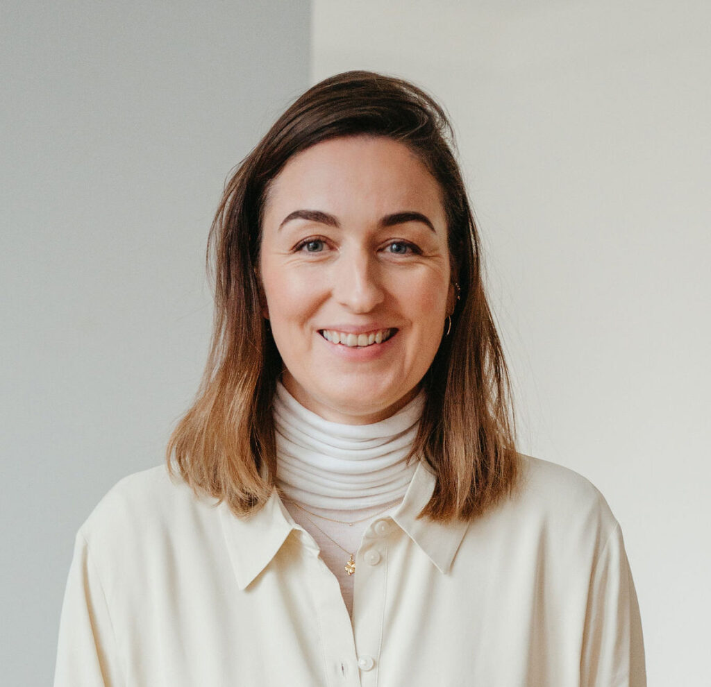 Nahaufnahme: Henrike in cremefarbener Bluse und weißem Rollkragen vor heller Wand.