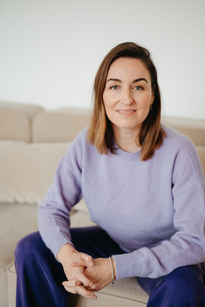 Nahaufnahme: Henrike Redecker im fliederfarbenen Pullover sitzt auf beigem Sofa und schaut in die Kamera.