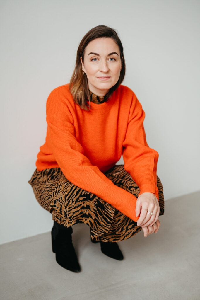 Henrike hockt vor heller Wand, orangefarbener Pullover, gemusterter Rock, schwarze Stiefel.