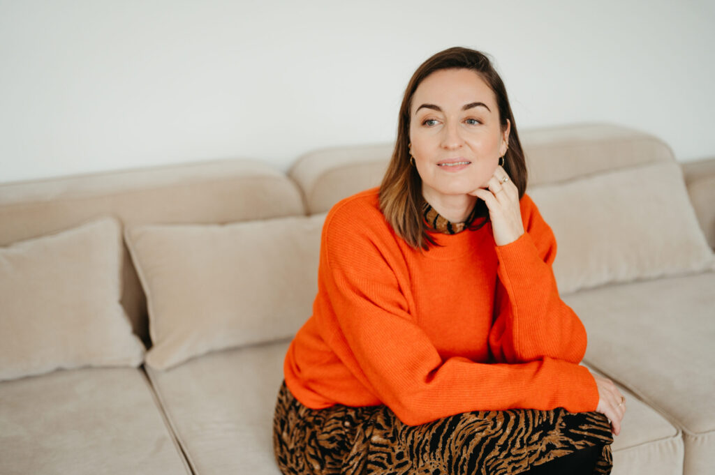 Henrike sitzt auf beigem Sofa, orangefarbener Pullover und gemusterter Rock; Hand am Kinn.