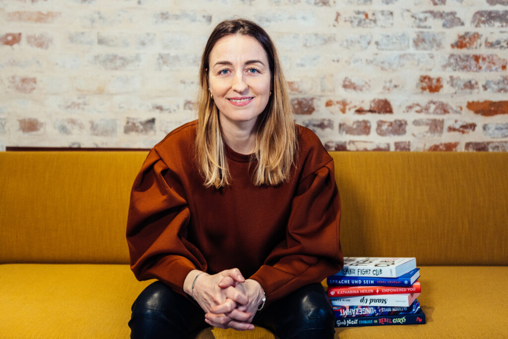 Henrike Redecker sitzt auf gelbem Sofa. Neben ihr liegt ein Stapel Bücher.